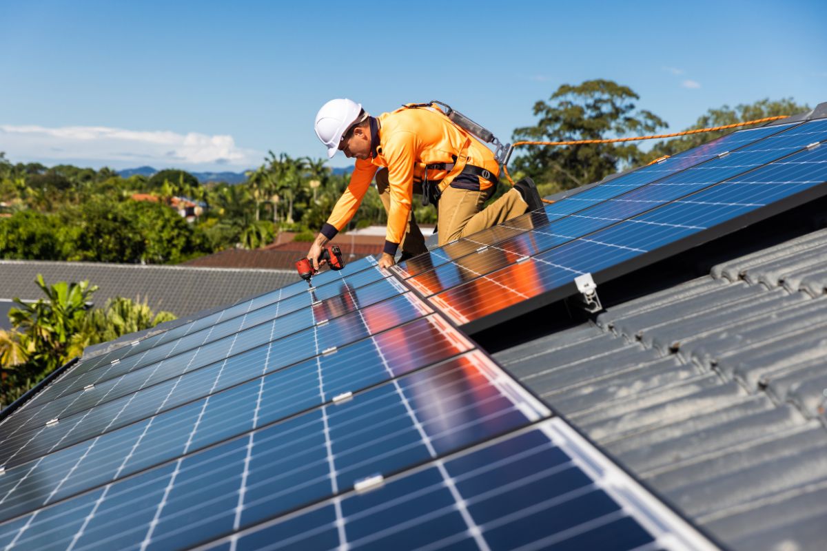 A man installing a solar panel on a rooftop, focused on his work under a clear blue sky.